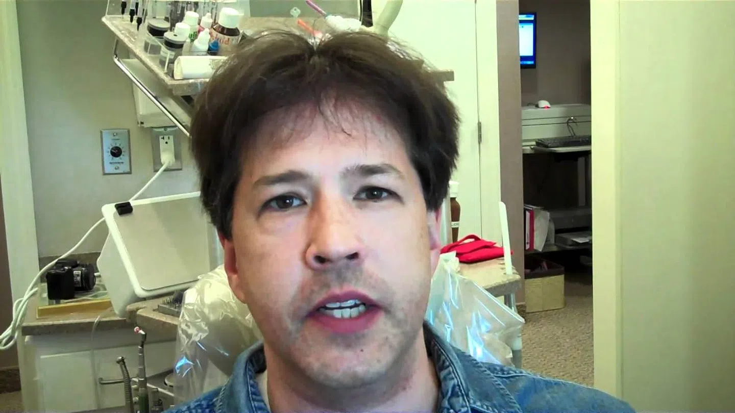 Man with brown hair speaking in a medical or dental office with equipment in the background