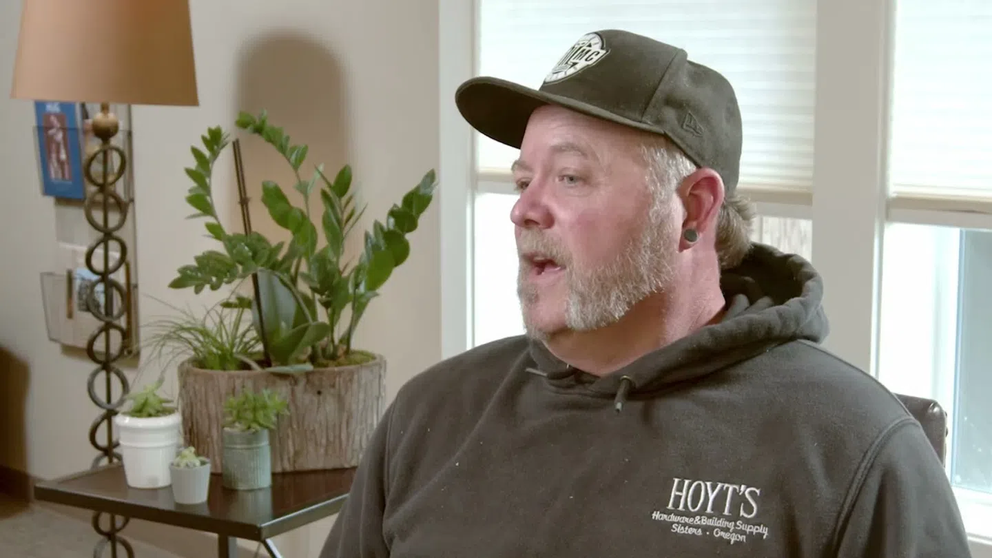 Man wearing black Hoyt's hoodie and cap sitting indoors with plants and lamps in the background
