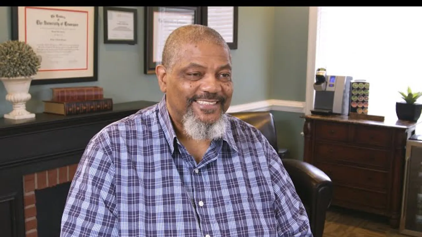 Smiling man with gray beard wearing a blue plaid shirt sitting in a cozy room with certificates and books behind.