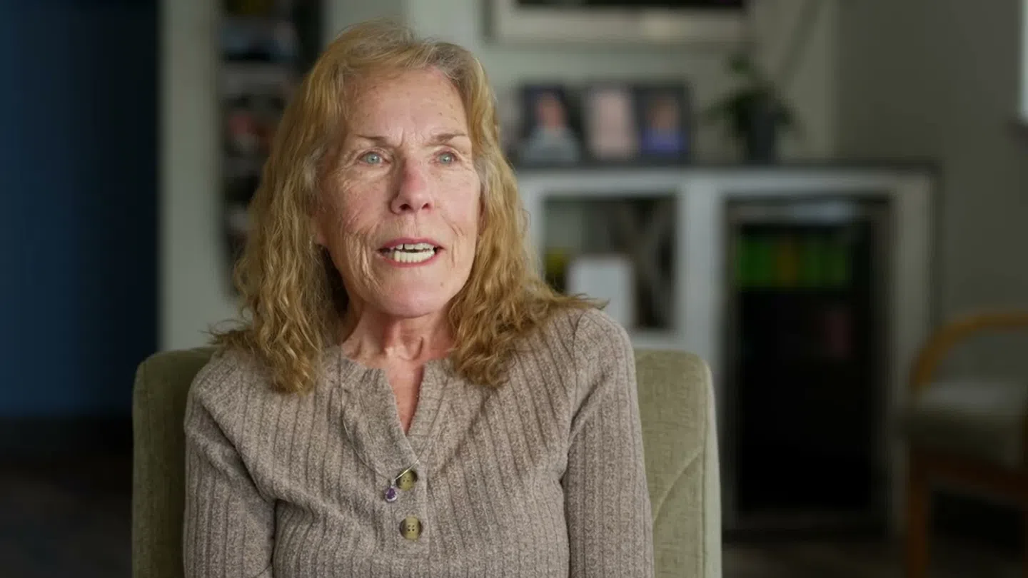 Older woman with curly blonde hair wearing a brown sweater sitting in a cozy room speaking.