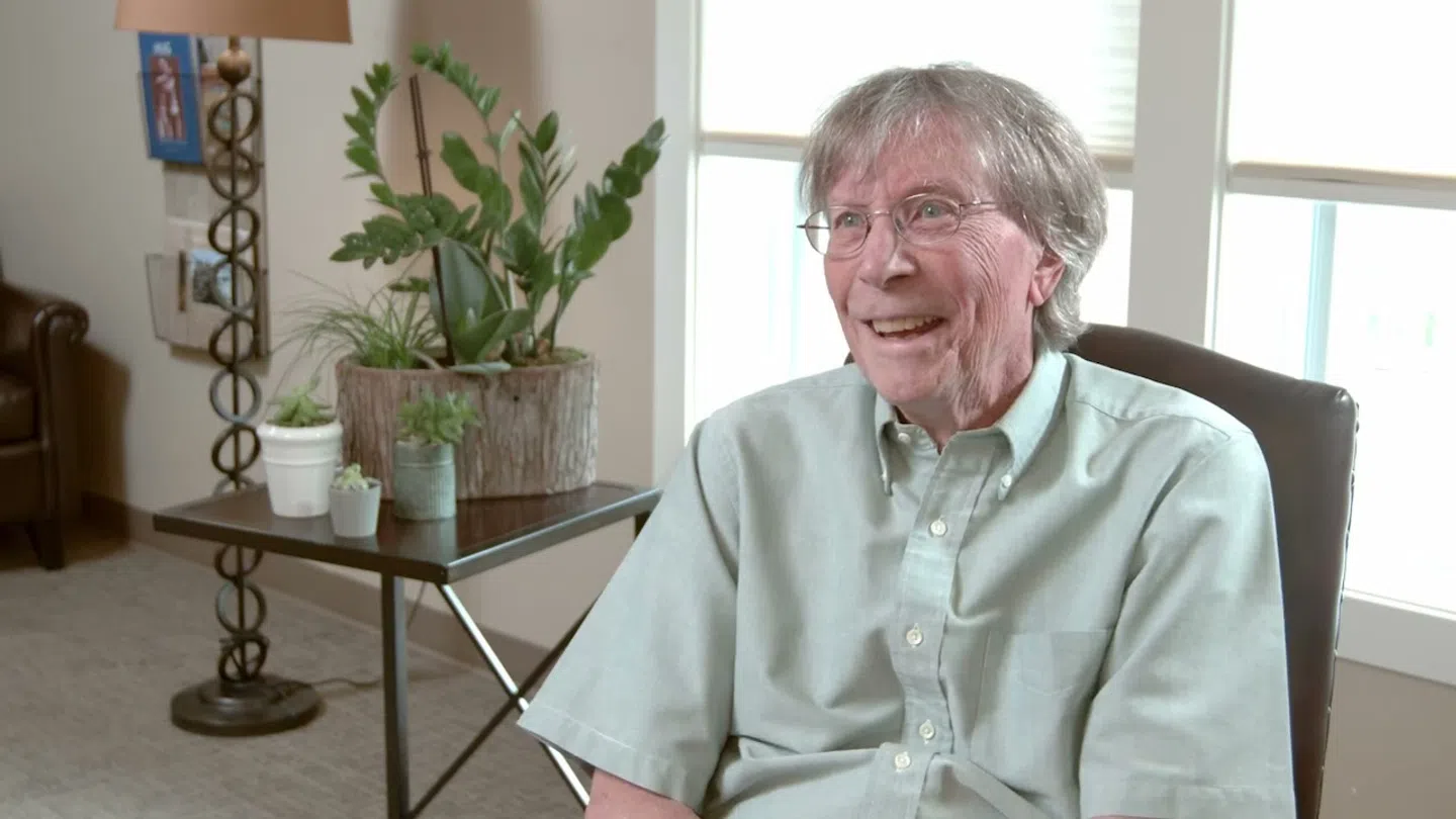 Elderly man smiling and seated in a bright room with indoor plants and a window in the background.