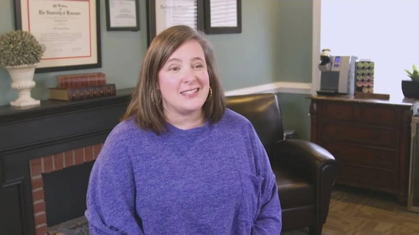 Woman in a purple sweater smiling while seated in a cozy office with framed certificates and a coffee machine.