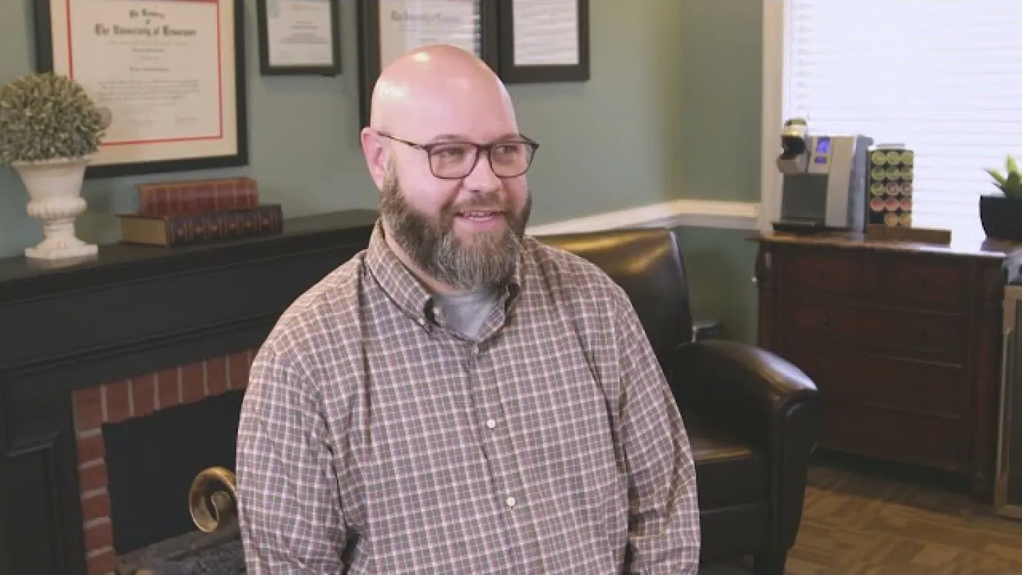 Smiling bearded man wearing glasses and plaid shirt sitting in cozy office with framed certificates and furniture.
