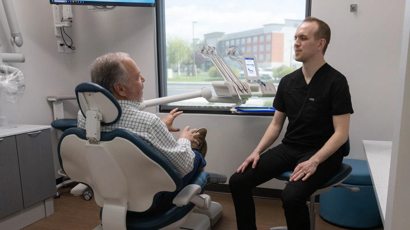 Dentist in black scrubs consulting an elderly male patient seated in a dental chair by a window.