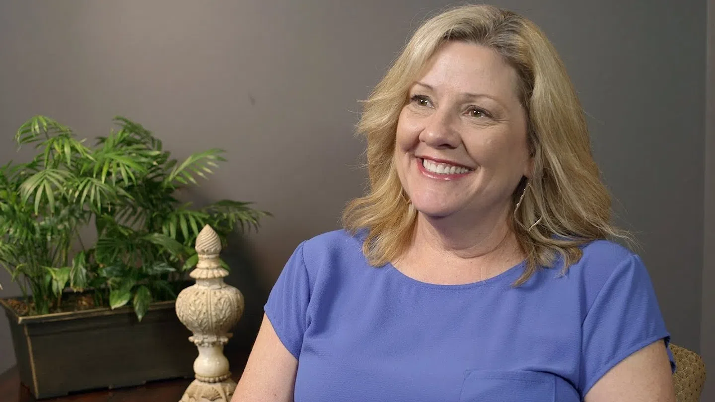 Smiling middle-aged woman with blonde hair wearing a blue shirt sitting indoors by a plant and decorative item.