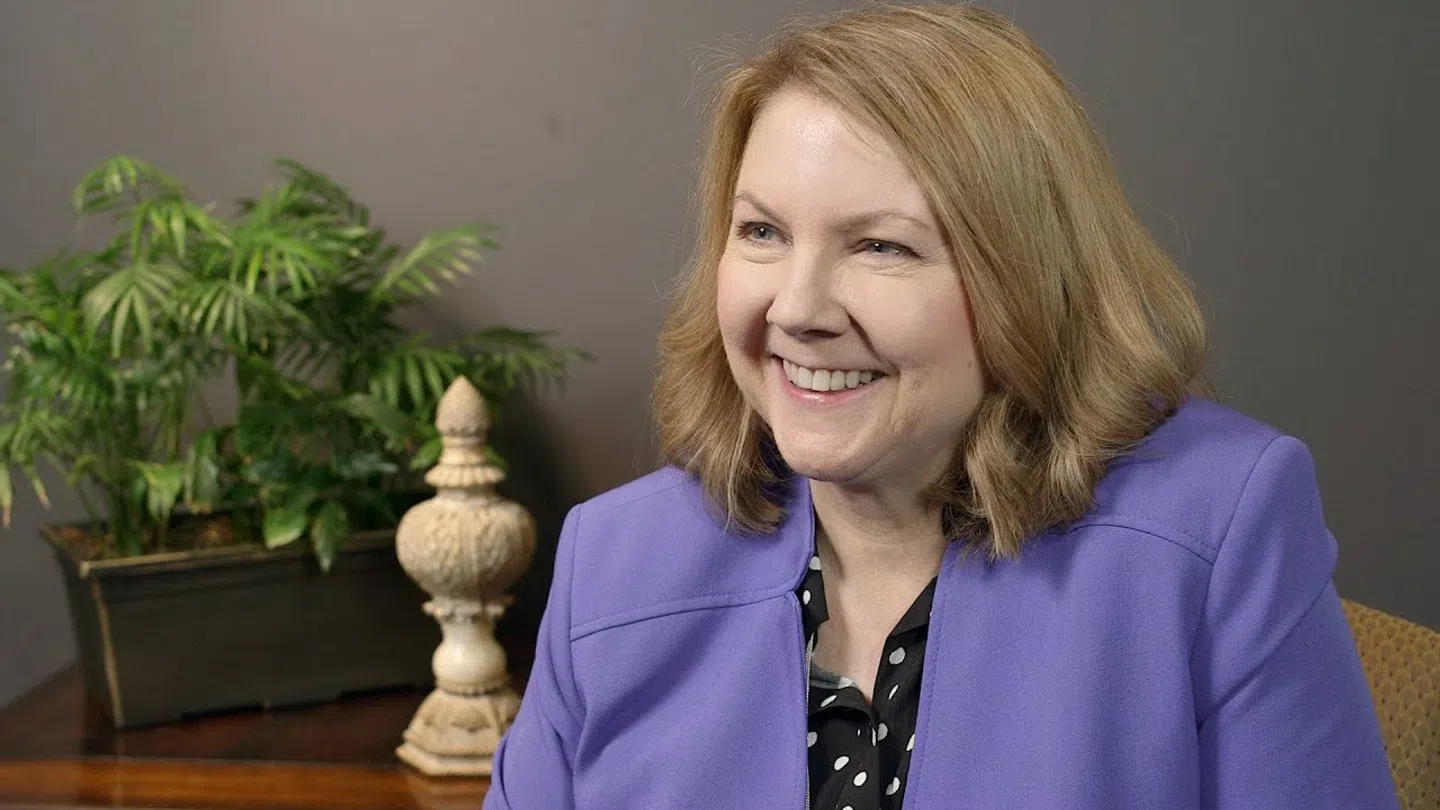 Smiling woman in a purple blazer sitting indoors with plants and decorative items in the background