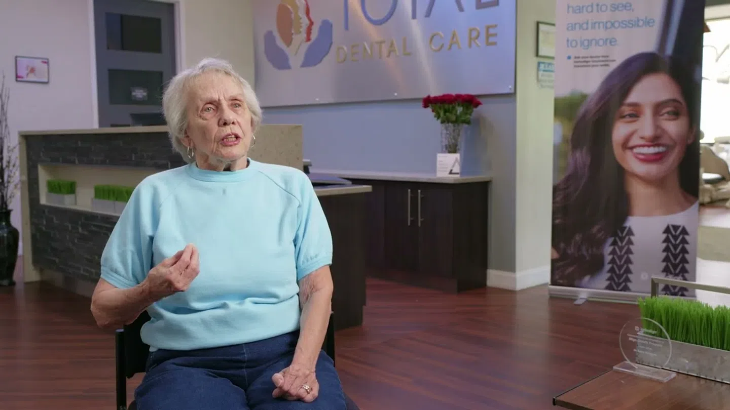 Elderly woman speaking in Total Dental Care office with dental signage and promotional banner in background