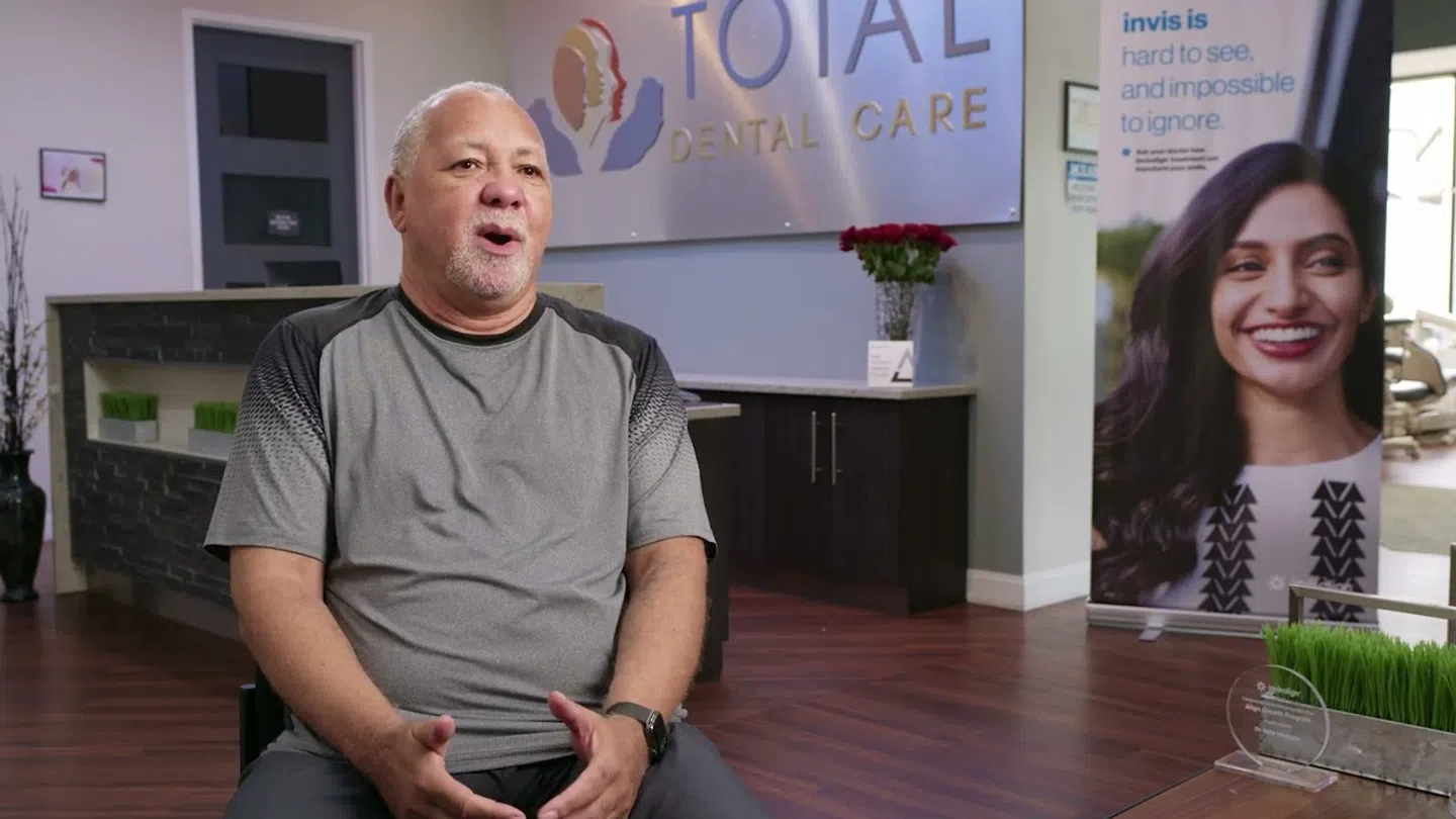 Man sitting and speaking inside a dental clinic with Total Dental Care sign and a smiling woman poster.