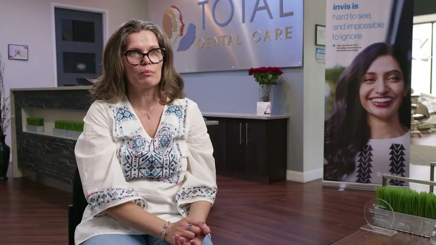 Middle-aged woman with glasses sitting in dental clinic Total Dental Care with promotional banner for Invisalign.