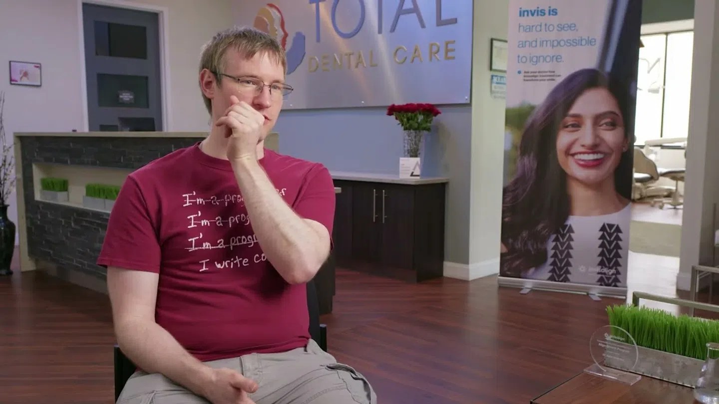Man in red shirt sitting in a Total Dental Care office with a smiling woman on an Invis banner in the background.