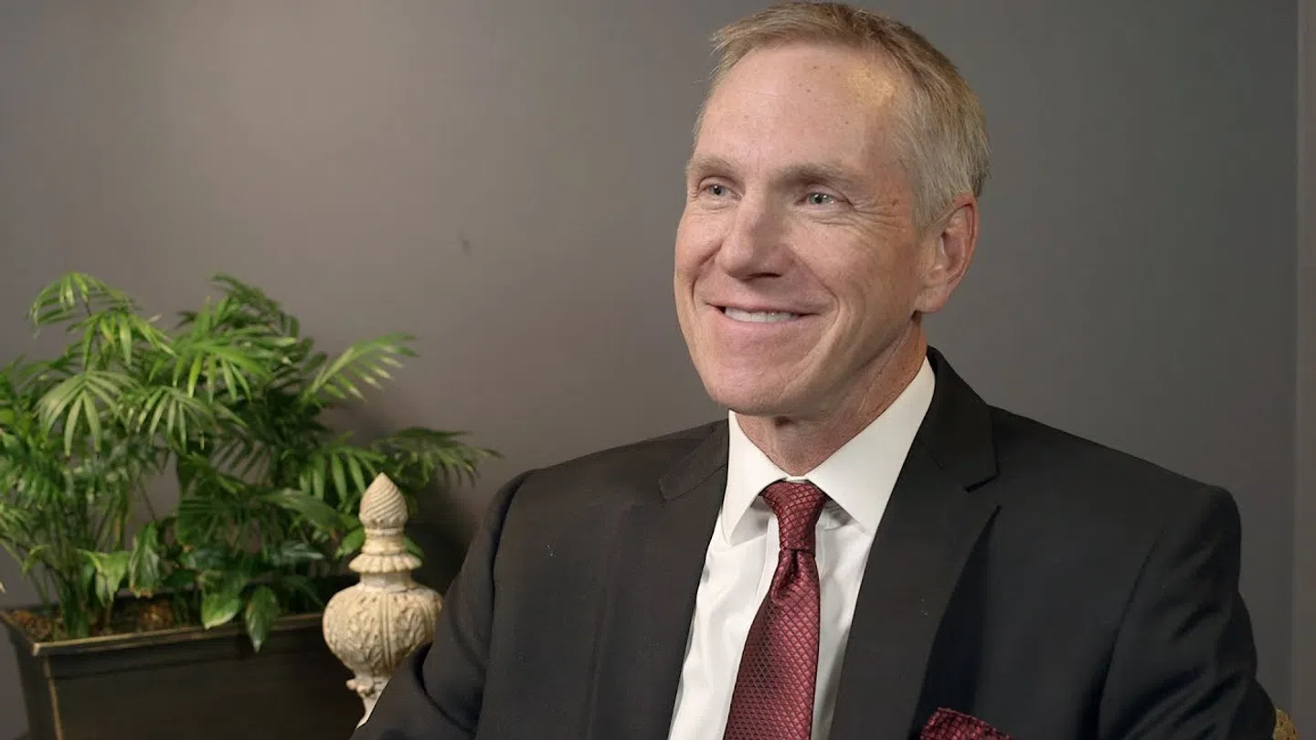 Smiling man in black suit and red tie seated indoors with green plant and dark gray wall background