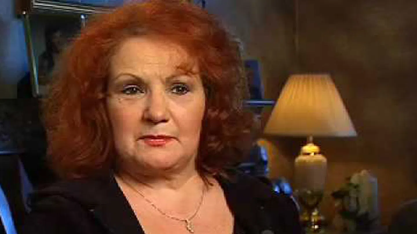Mature woman with red curly hair wearing a necklace, sitting indoors next to a lit table lamp in a cozy room.