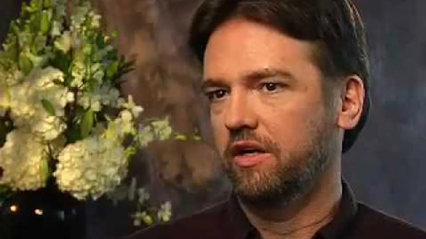 Man with beard speaking in an interview setting with a floral arrangement in the blurred background