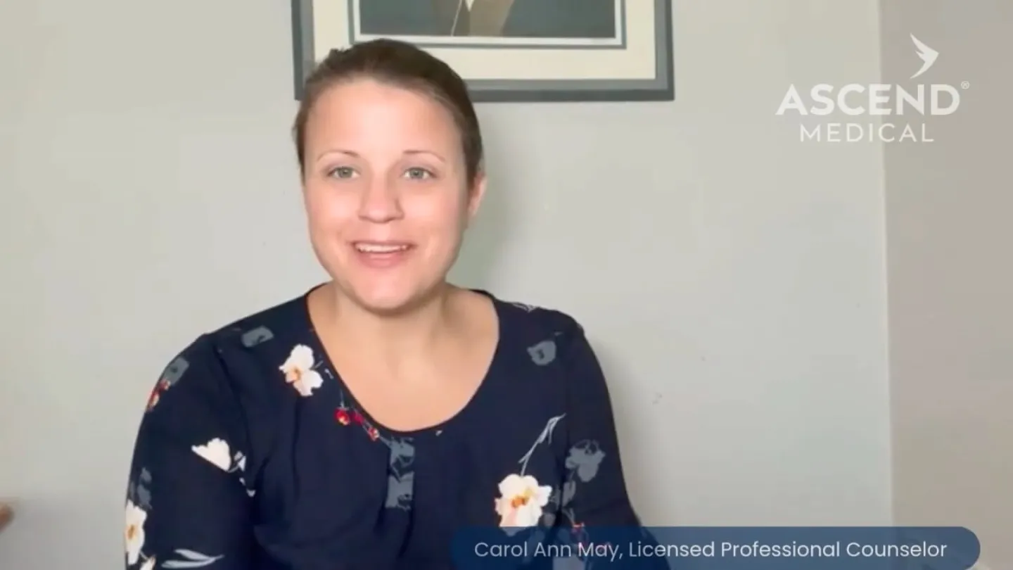 Smiling female licensed professional counselor in floral top sitting indoors with Ascend Medical logo in background.