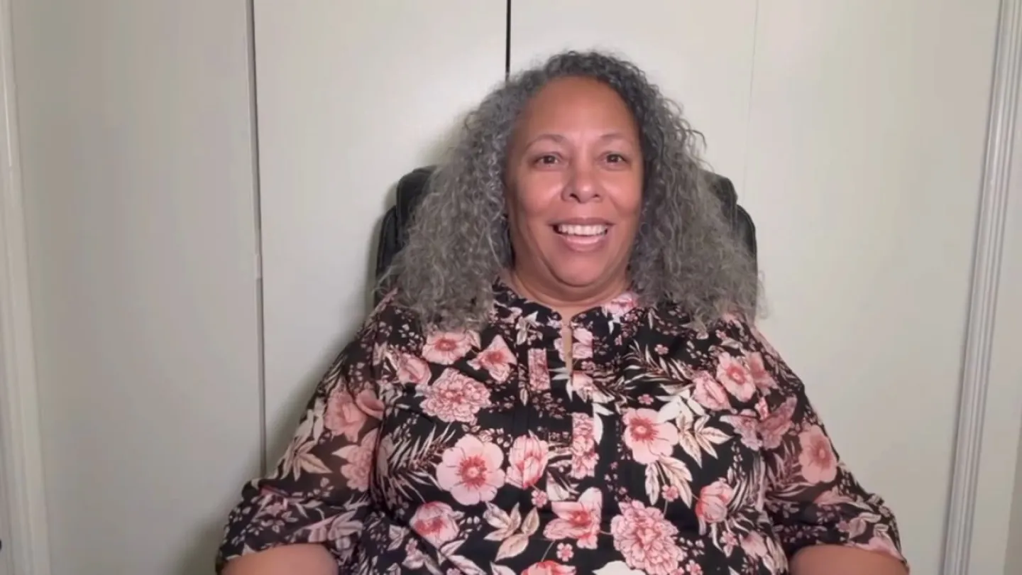 Smiling woman with curly gray hair wearing a floral blouse sitting against a plain wall background
