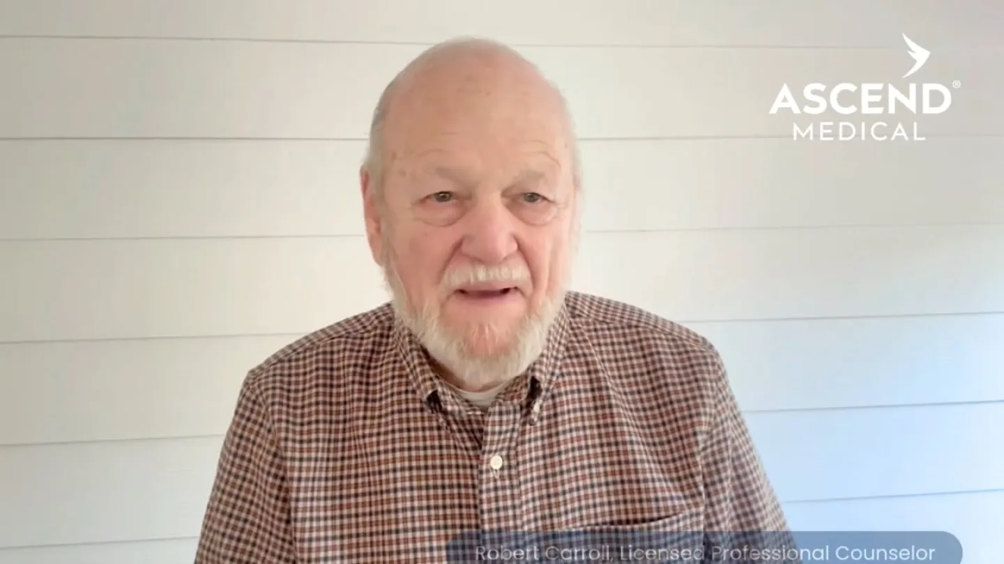 Elderly man in checkered shirt speaking in front of white shiplap wall with Ascend Medical logo