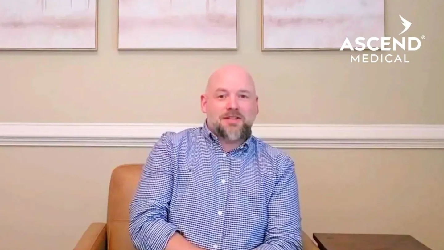 Man in blue checkered shirt sitting in a chair with Ascend Medical logo in background