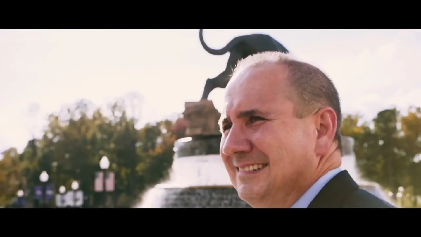 Smiling man in a suit near a water fountain with a black panther statue and trees in the background.
