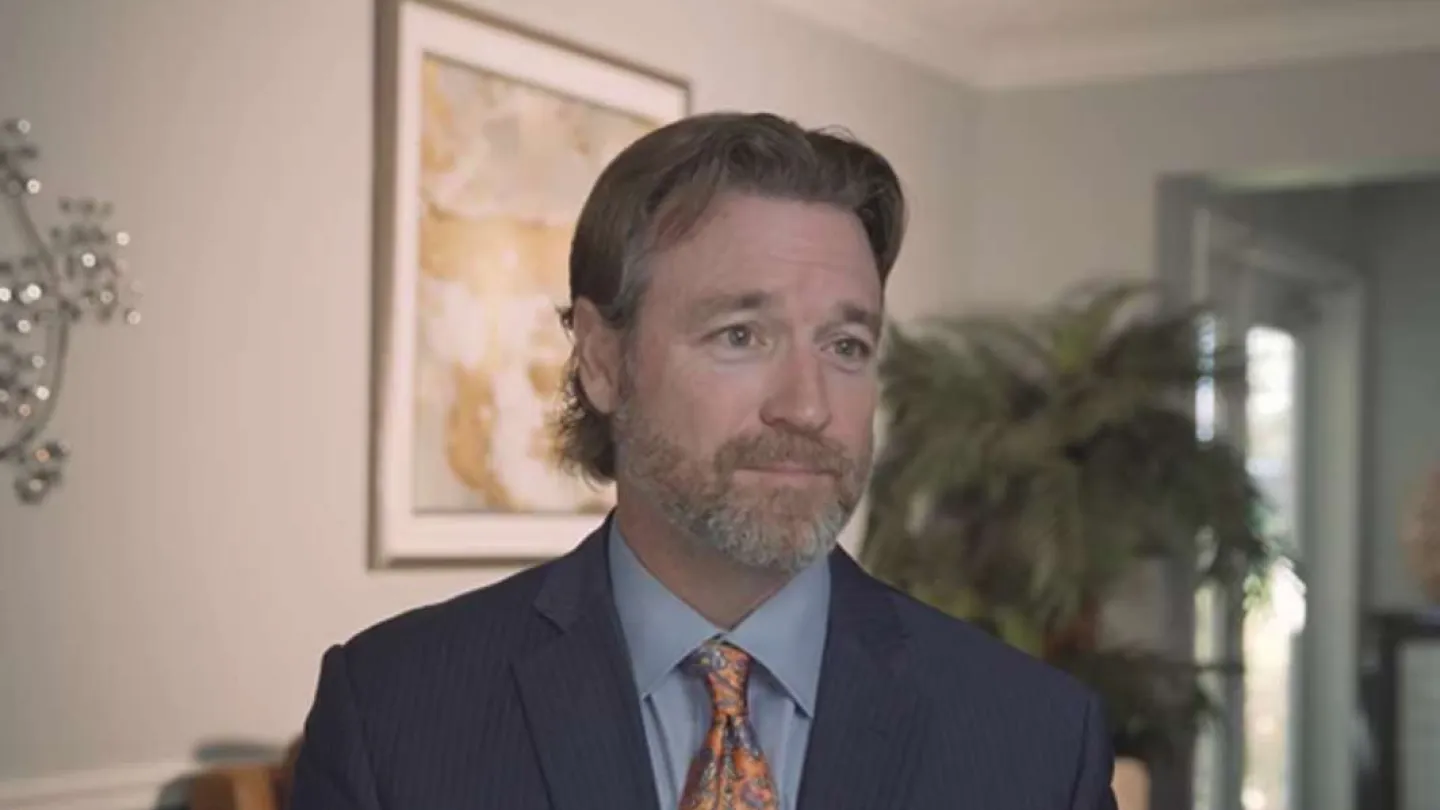 Man in a suit and patterned tie standing in a modern room with artwork and plants in the background