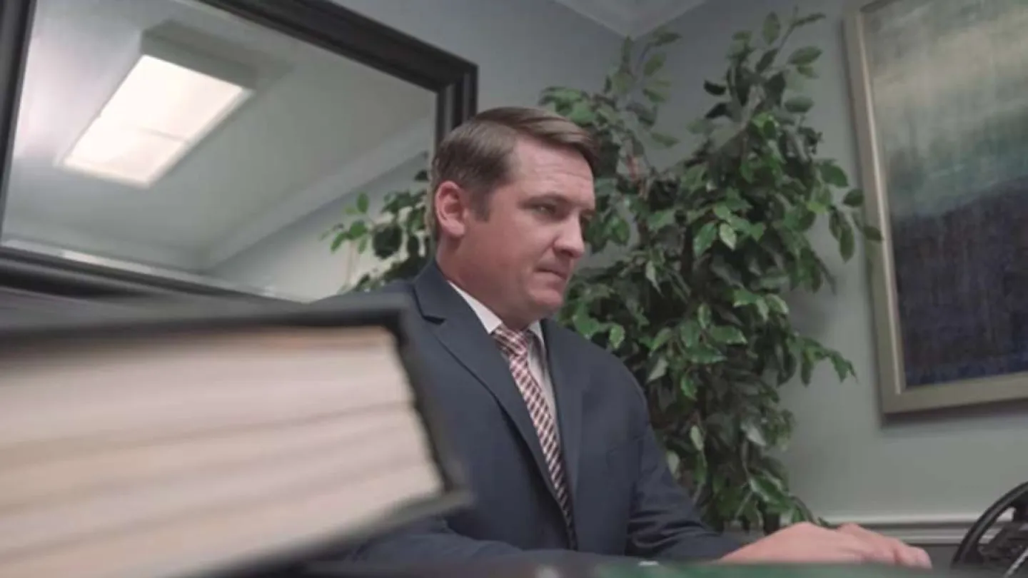 Man in suit working at desk with large book in foreground and plant in office background