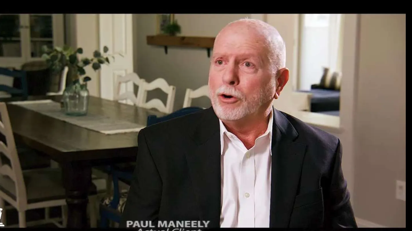 Elderly man in black suit and white shirt speaking in a well-lit dining room with wooden table and chairs.