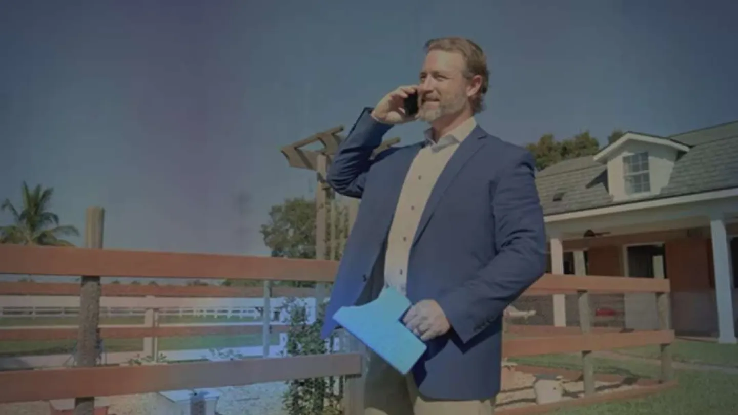 Man in blue blazer talking on phone outside house next to wooden fence on sunny day