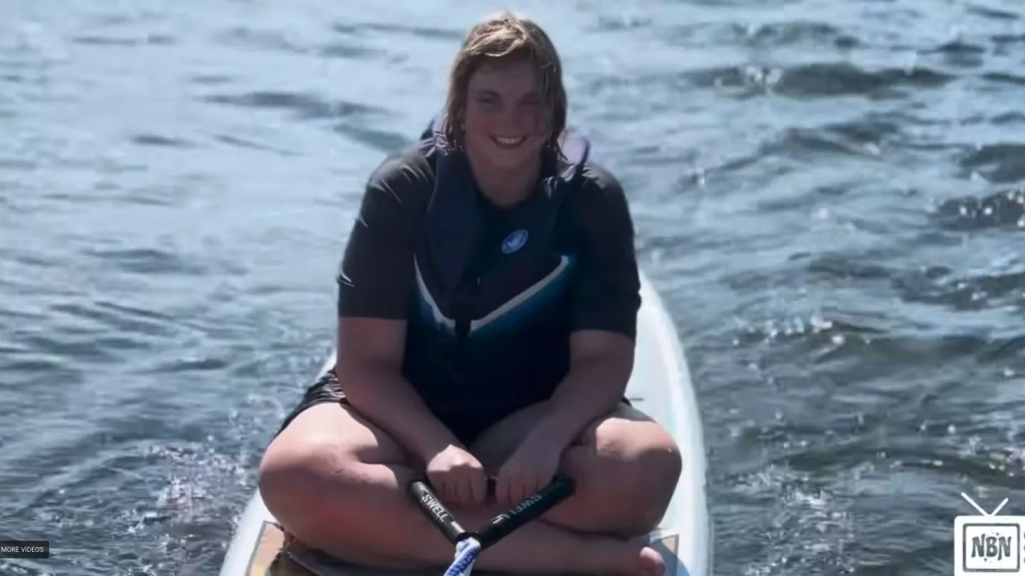 Smiling person in black wetsuit sitting cross-legged on paddleboard on calm water under sunlight.