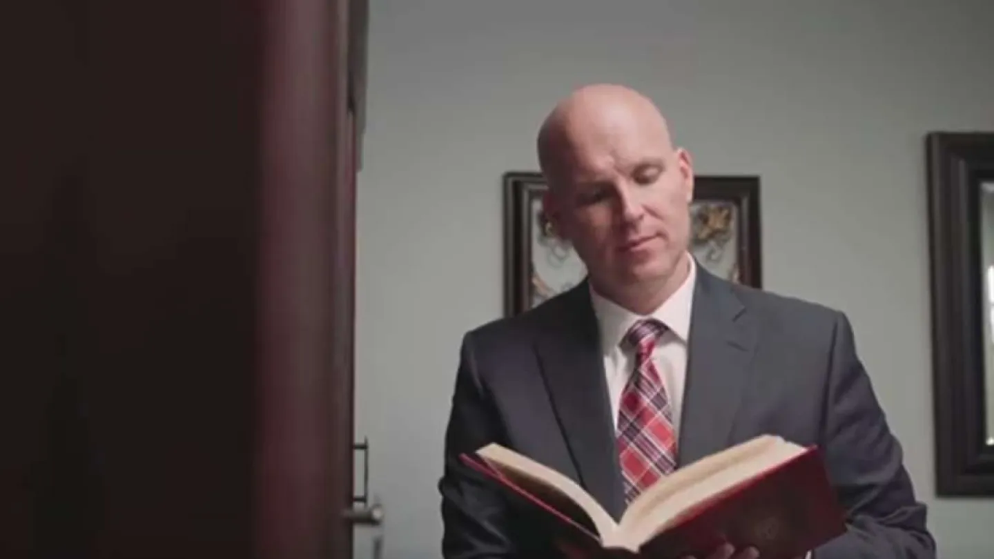Bald man in a suit and plaid tie reading a large red book indoors with framed pictures on the wall.