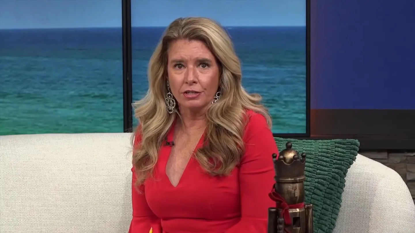 Woman in a red dress with long blonde hair sitting on a white couch with ocean view behind her.