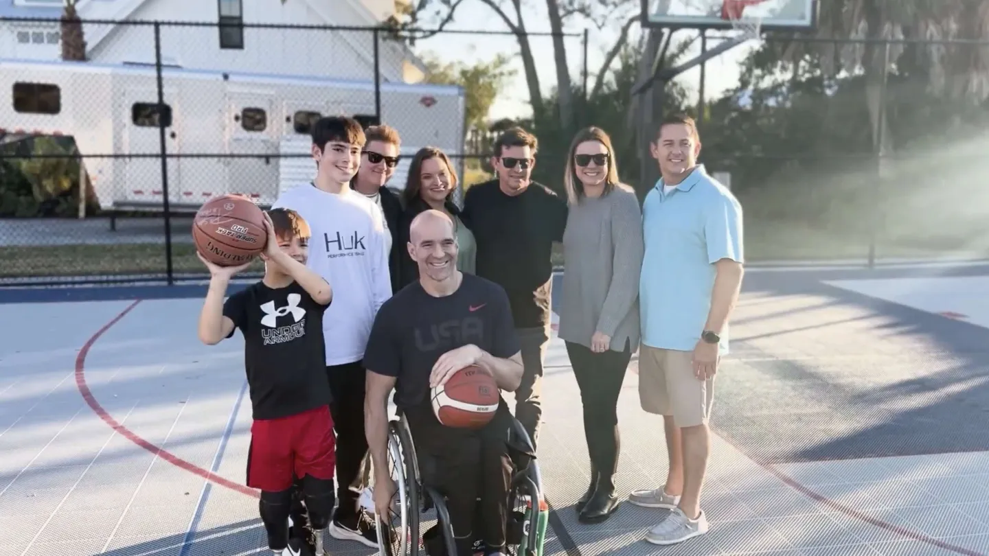 A diverse group of smiling people, including a man in a wheelchair holding a basketball, on an outdoor basketball court.