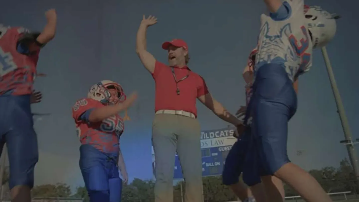 Football coach celebrating with young players during a daytime outdoor practice or game.