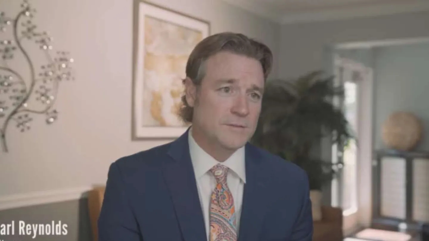 Man in a blue suit and colorful tie speaking in a well-lit modern office with wall art and plants.