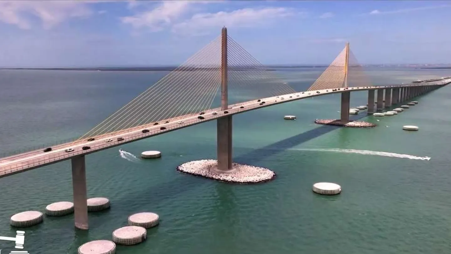 Aerial view of a modern cable-stayed bridge over turquoise water with vehicles and boats under a blue sky.