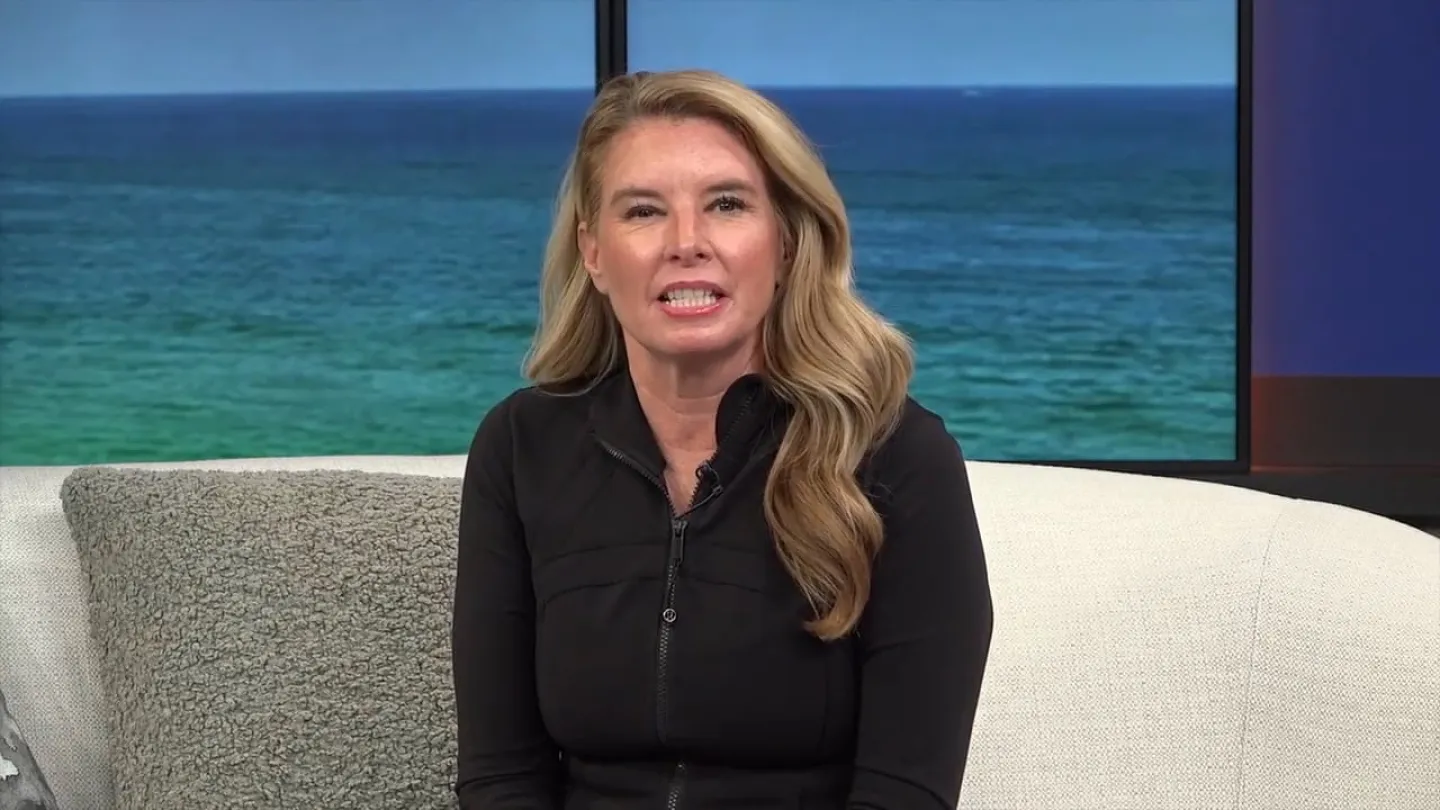 Woman with long blonde hair in black jacket sitting on a couch with ocean view behind her