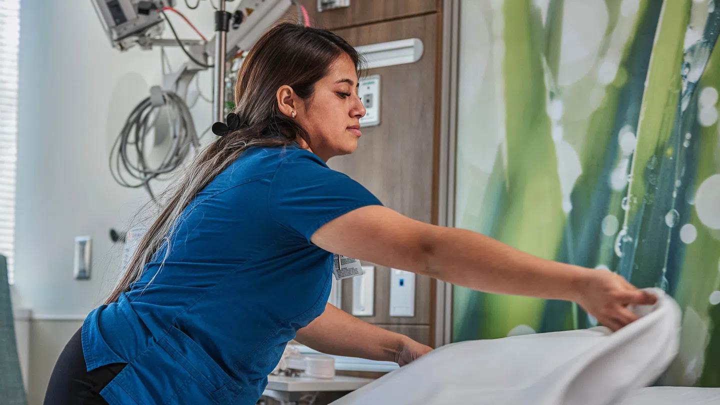 Healthcare worker in blue scrubs making a hospital bed with white linens in a patient room.