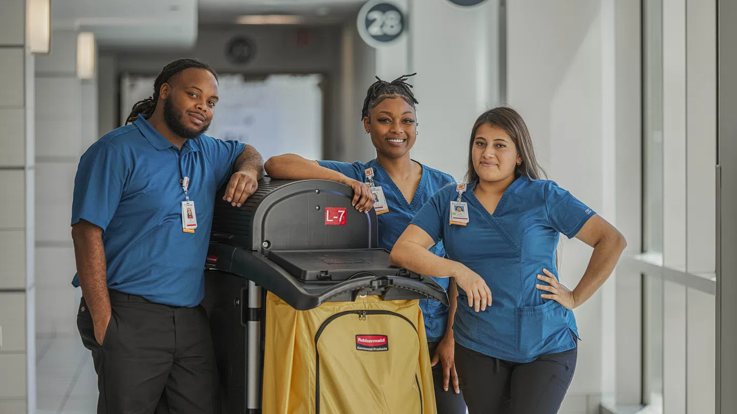 Three janitorial staff in blue uniforms pose with a yellow cleaning cart in a bright hallway.