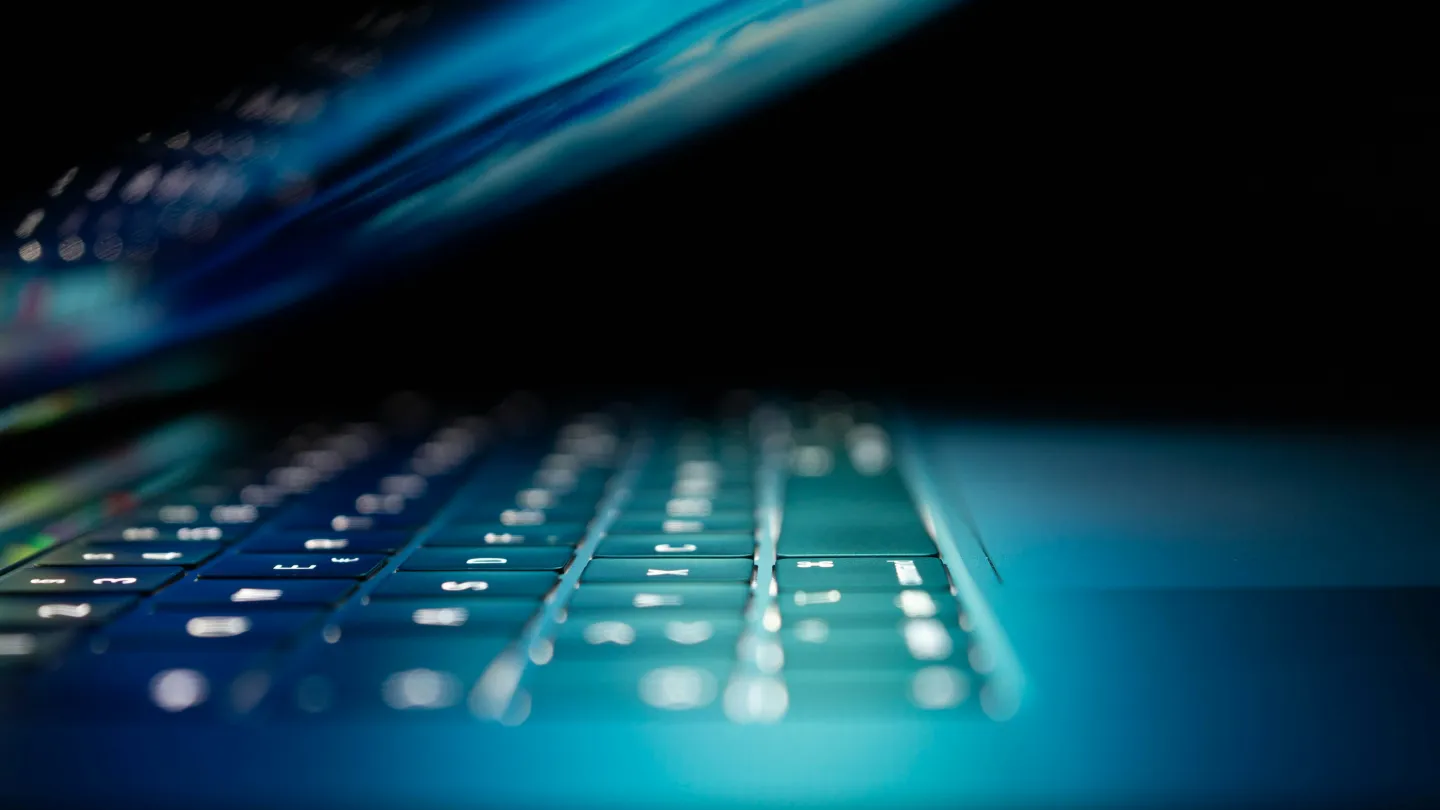 Close-up of a backlit laptop keyboard with glowing keys and partially open screen in low light.
