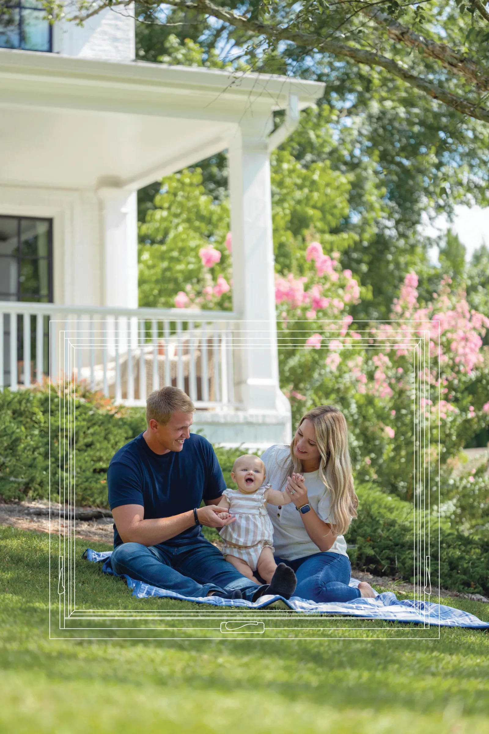 Happy family sitting on a blanket in the garden outside a white house with blooming pink flowers.