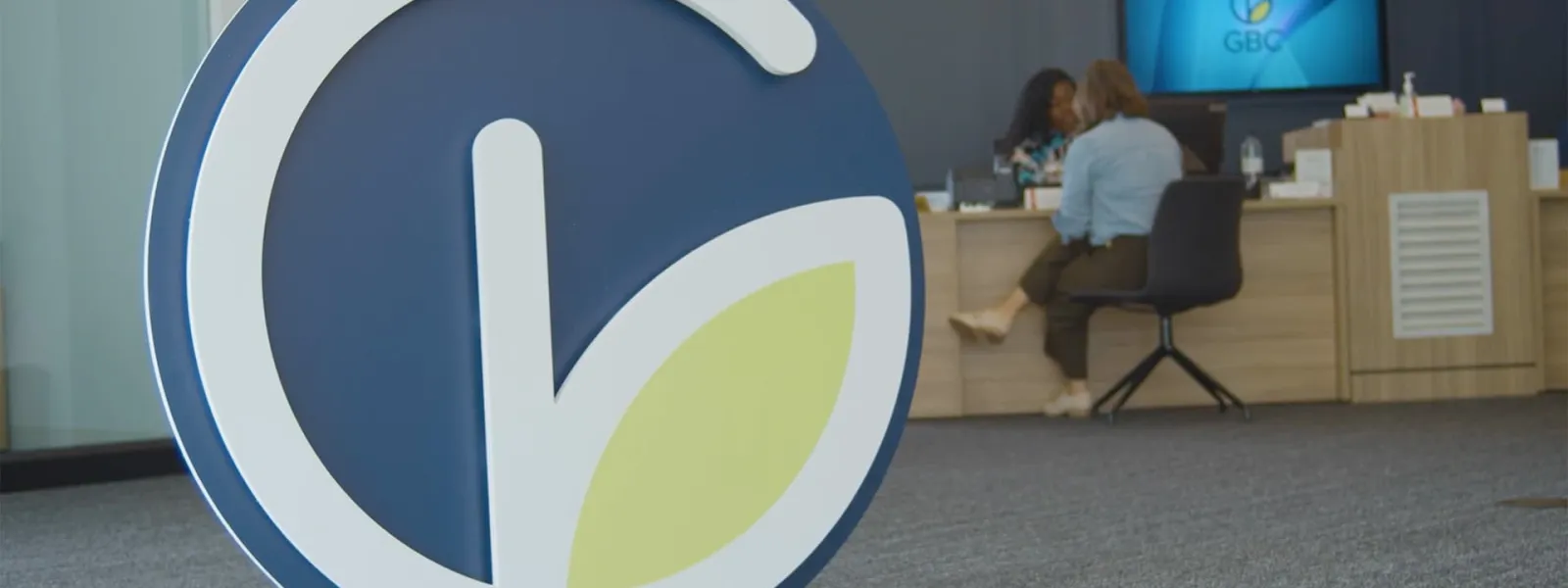 Large GBC logo with a leaf design in office lobby where two people are seated at a wooden desk in the background