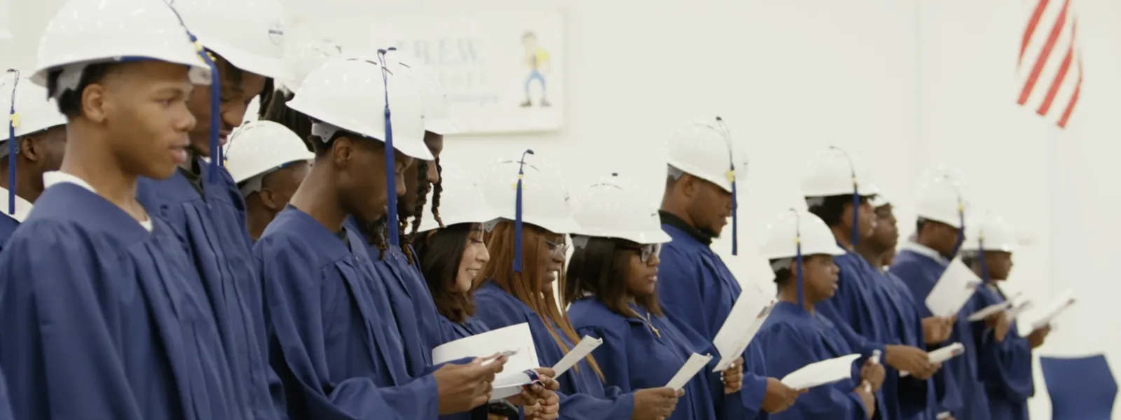 Graduates in blue gowns and white hard hats reading from papers during a ceremony indoors