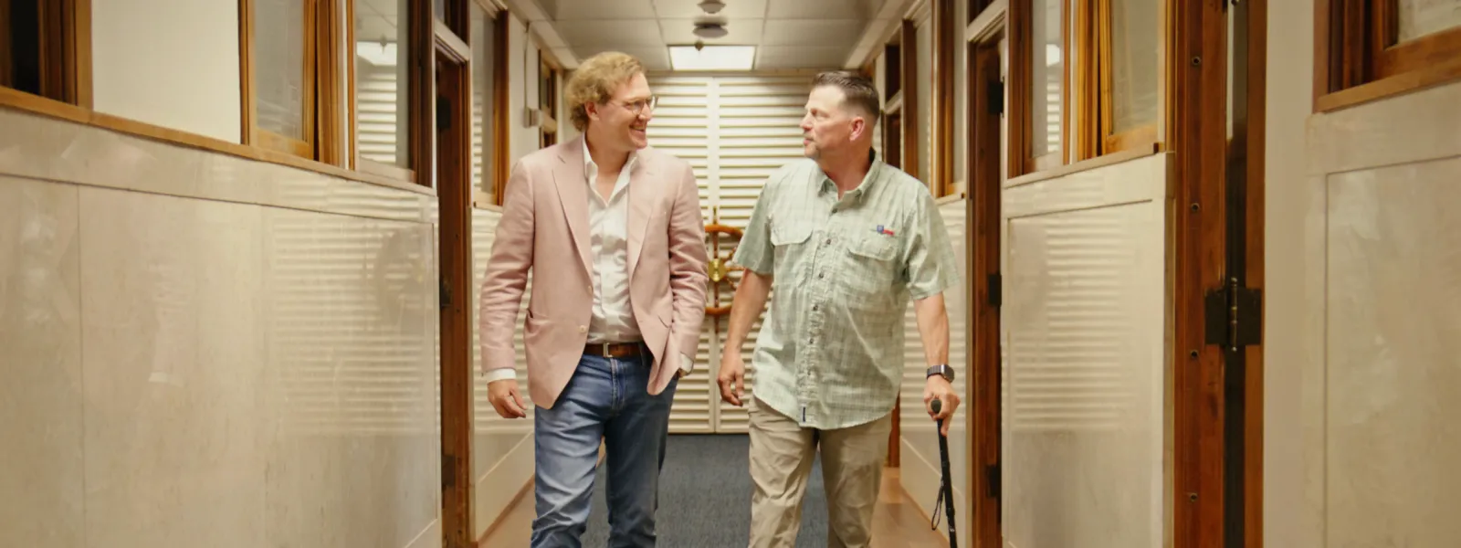 Two men walking and talking in a brightly lit office hallway with wooden doors and blue carpet.
