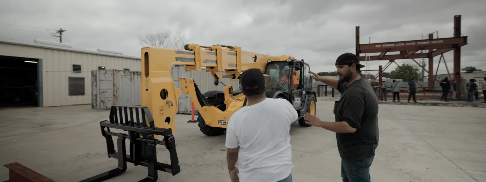 Two workers inspecting a yellow telehandler forklift in an outdoor industrial training area under cloudy sky