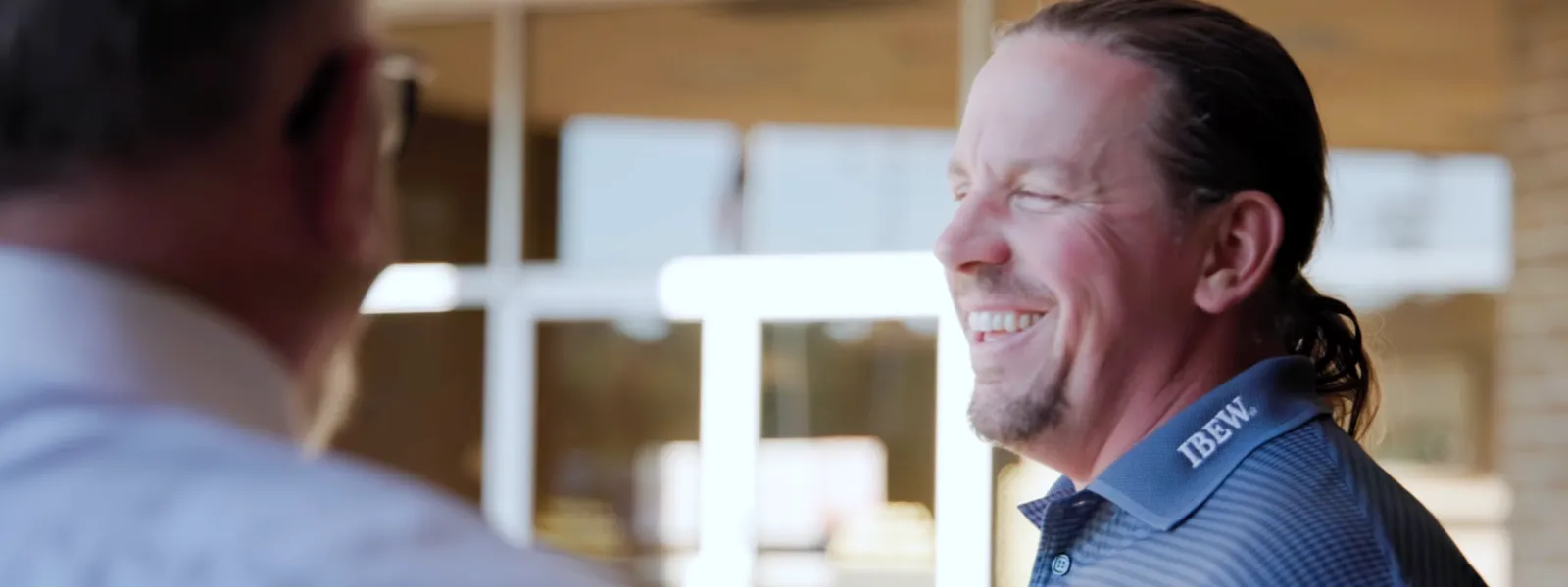 Man in blue IBEW polo shirt smiling and talking with another man in a professional setting near glass doors.