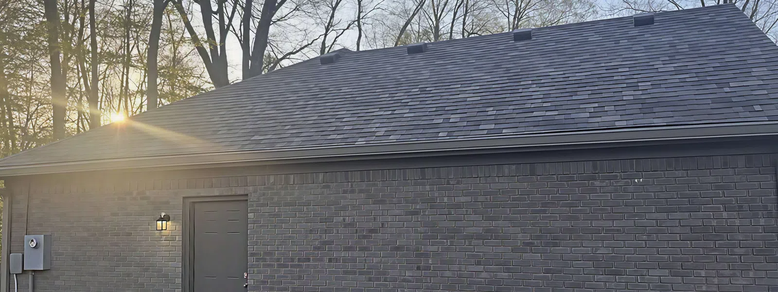 Aerial view of a house roof with gray asphalt shingles next to a green lawn and neighboring brick house..