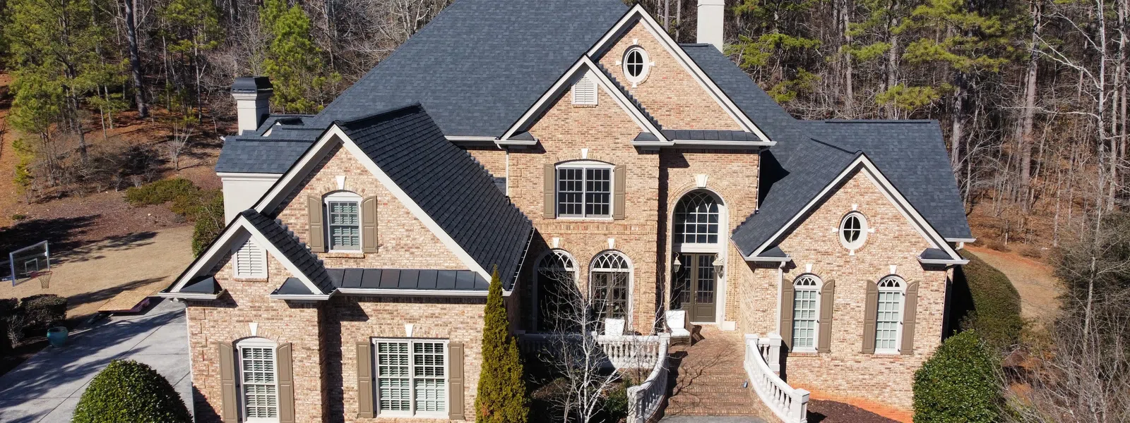 Large brick house with multiple gables, arched windows, slate residential roofing, surrounded by trees and manicured bushes..