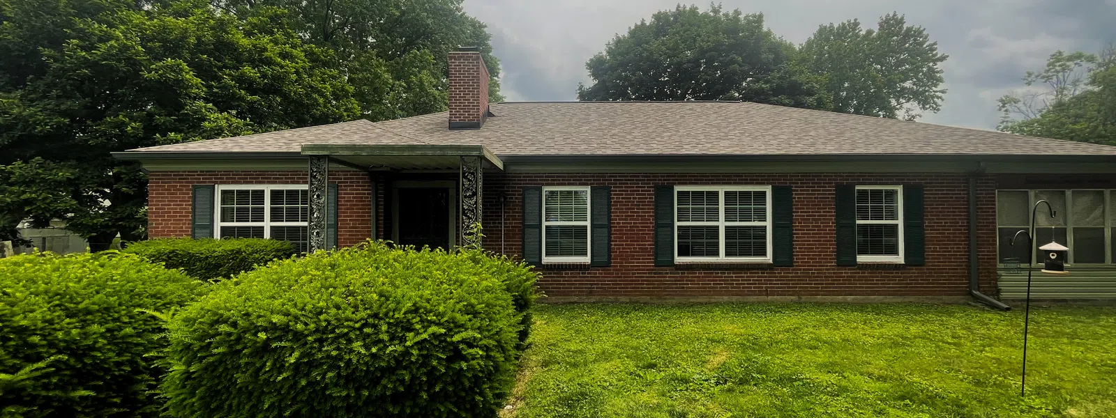 Single-story brick house with black shutters, asphalt residential roofing, manicured bushes, and cloudy sky overhead in a green yard..