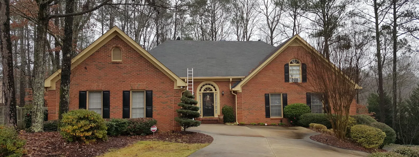 Beautiful brick home with asphalt residential roofing, surrounded by lush greenery and trees, featuring a well-maintained driveway and landscaping..