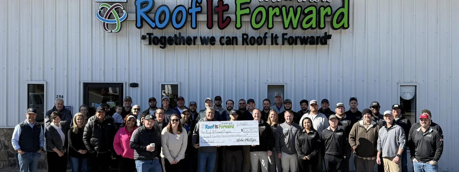 Large group of people posing outside building with Roof It Forward sign holding oversized check for donation..