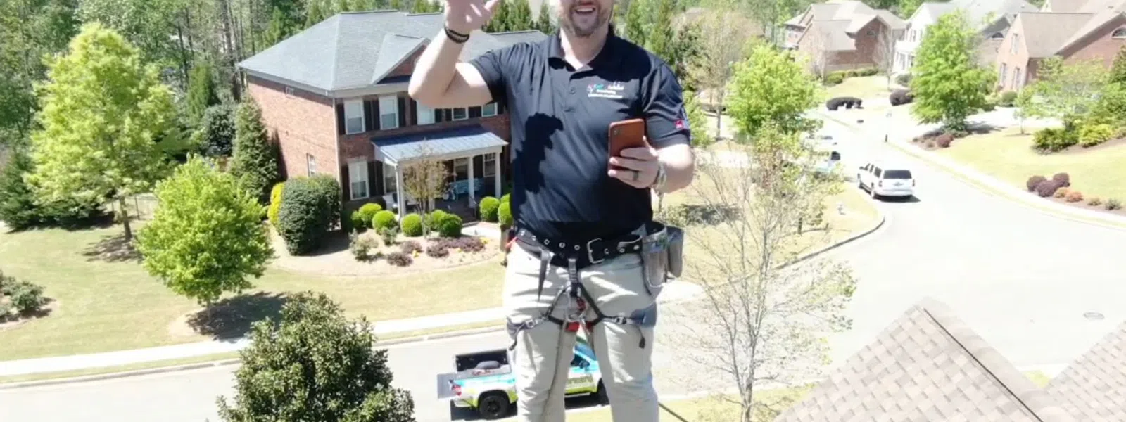 A smiling roofer on a house rooftop, holding a phone and waving to the camera in a residential neighborhood..