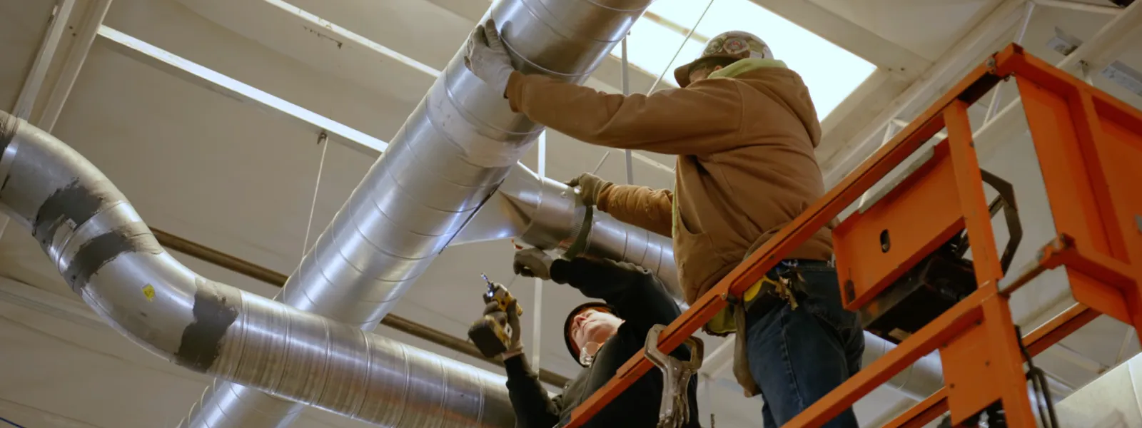 Two workers install large silver ventilation ducts inside an industrial building from an elevated platform.